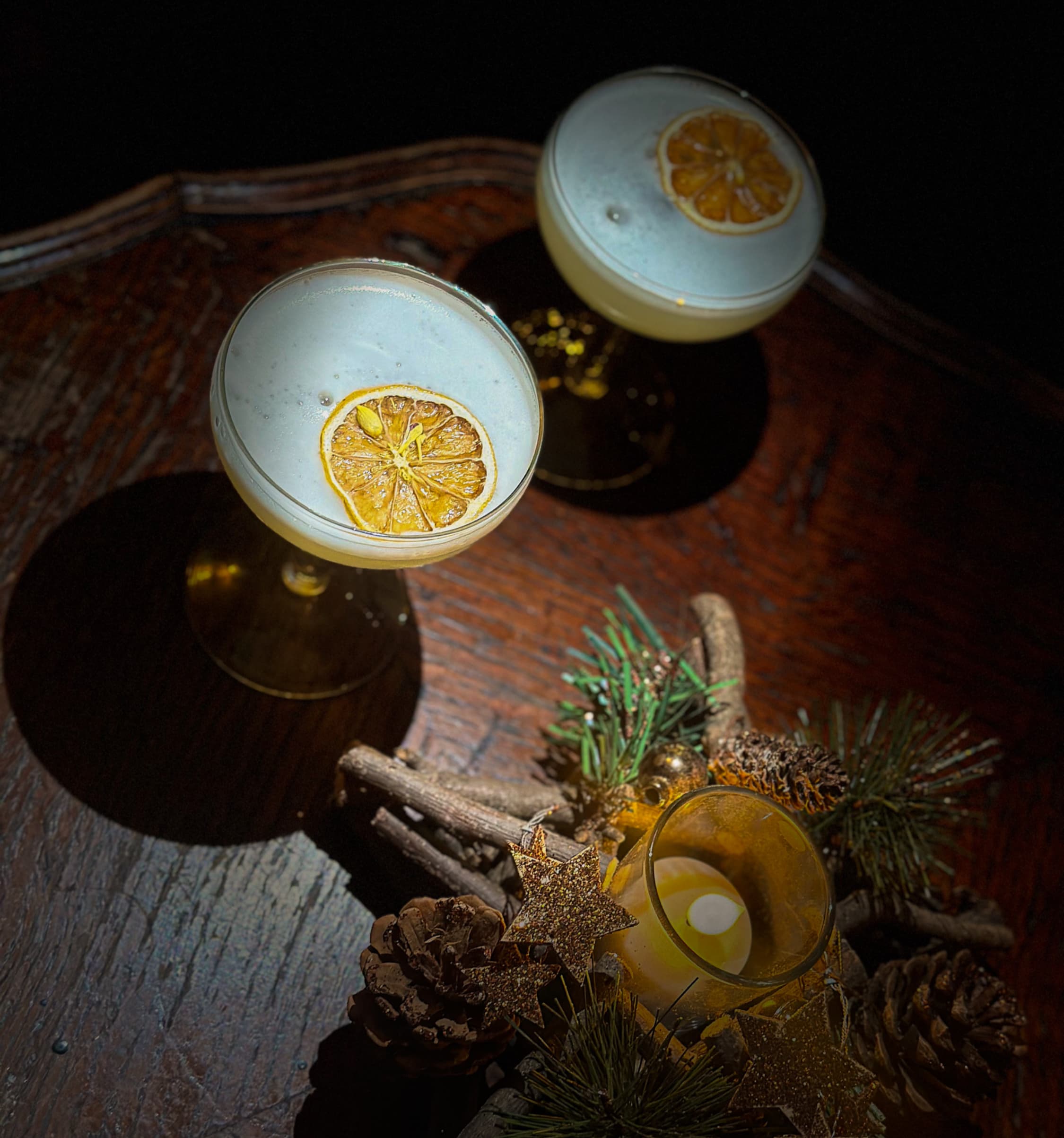 Two foamed coupe cocktails crowned with dried citrus, side-lit on a dark tray.